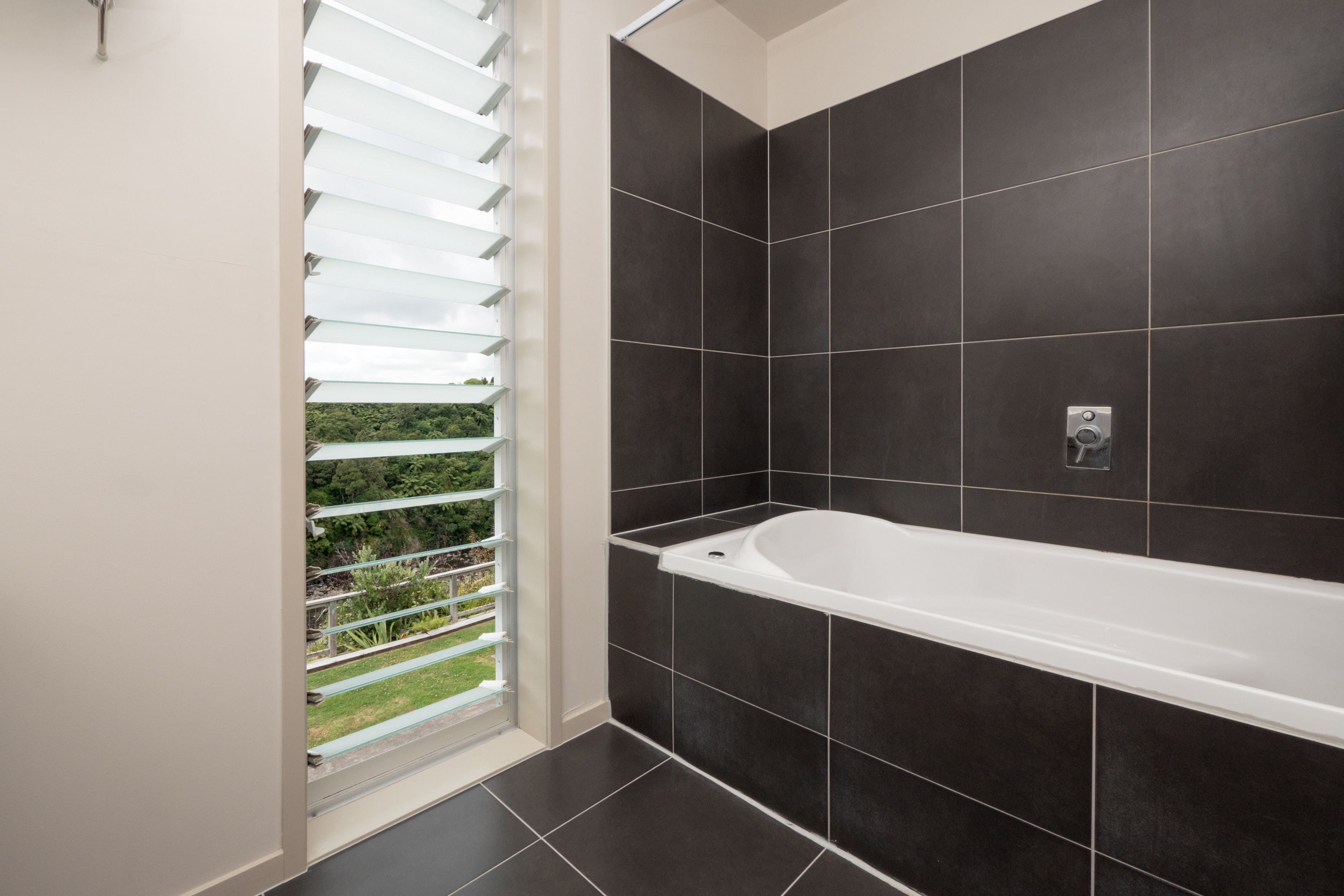 A modern bathroom with dark tiles, a white bathtub, and a window with blinds.