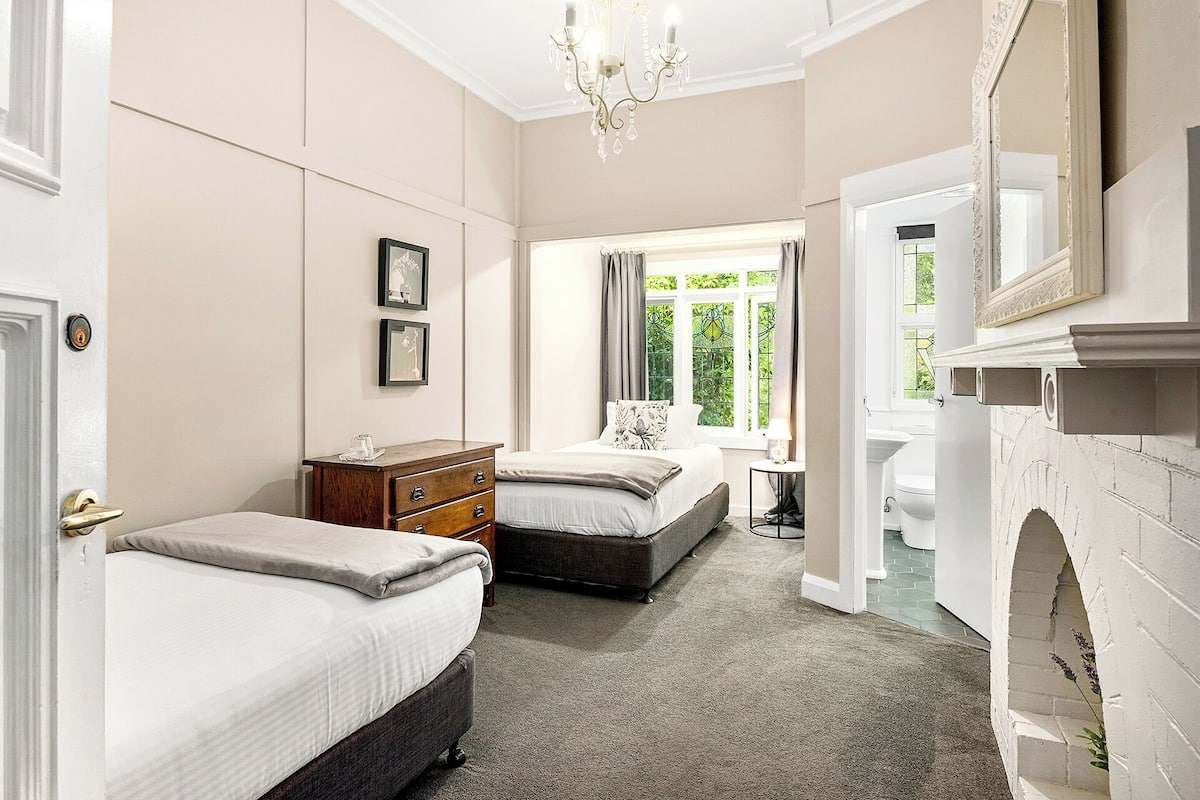 A bedroom with two beds, a dresser, a desk, a chandelier, and a fireplace.