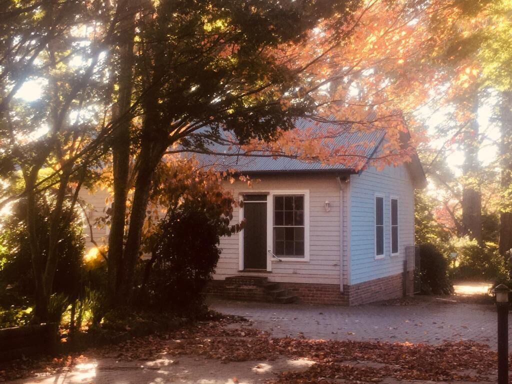 Une petite maison à un étage, avec une porte d’entrée et des fenêtres, entourée d’arbres aux feuilles d’automne.