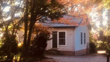 Une petite maison à un étage, avec une porte d’entrée et des fenêtres, entourée d’arbres aux feuilles d’automne.