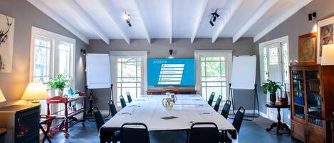 Un salón de conferencias con una larga mesa, sillas, una pantalla de proyector que muestra un orden del día y ventanas con persianas.