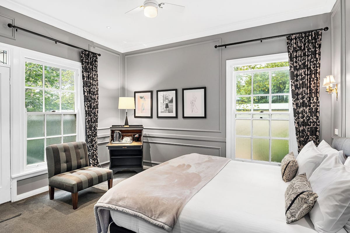 A hotel room with a large bed, a chair, a small table with a lamp, and two windows with curtains.