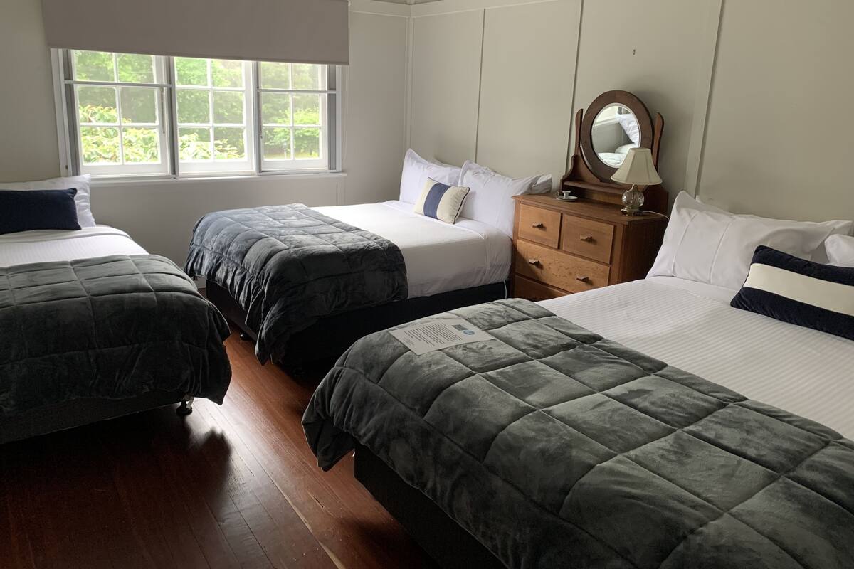 A hotel room with three beds, a wooden dresser, a mirror, and a window with blinds.