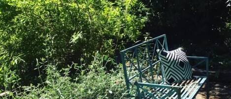 A green metal outdoor seating area with a round table and a bench, surrounded by dense foliage.