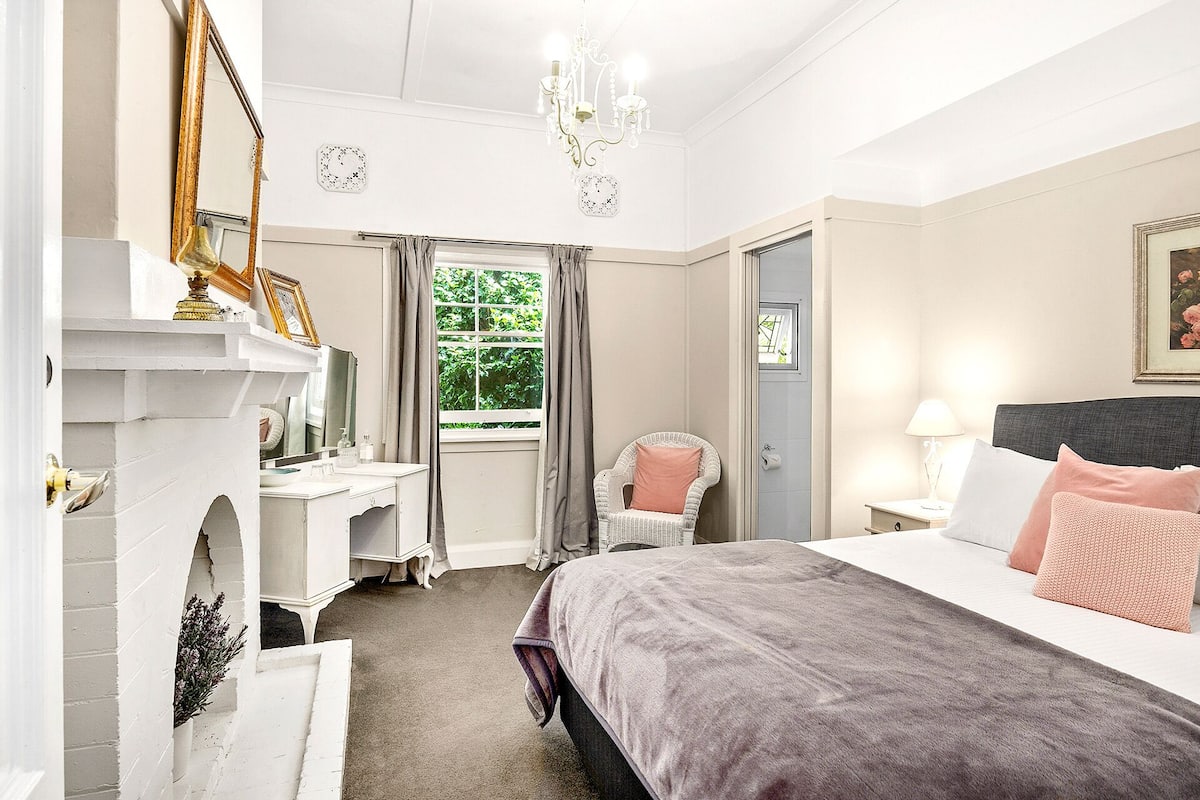 A bedroom with a bed, a chair, a desk, and a fireplace.