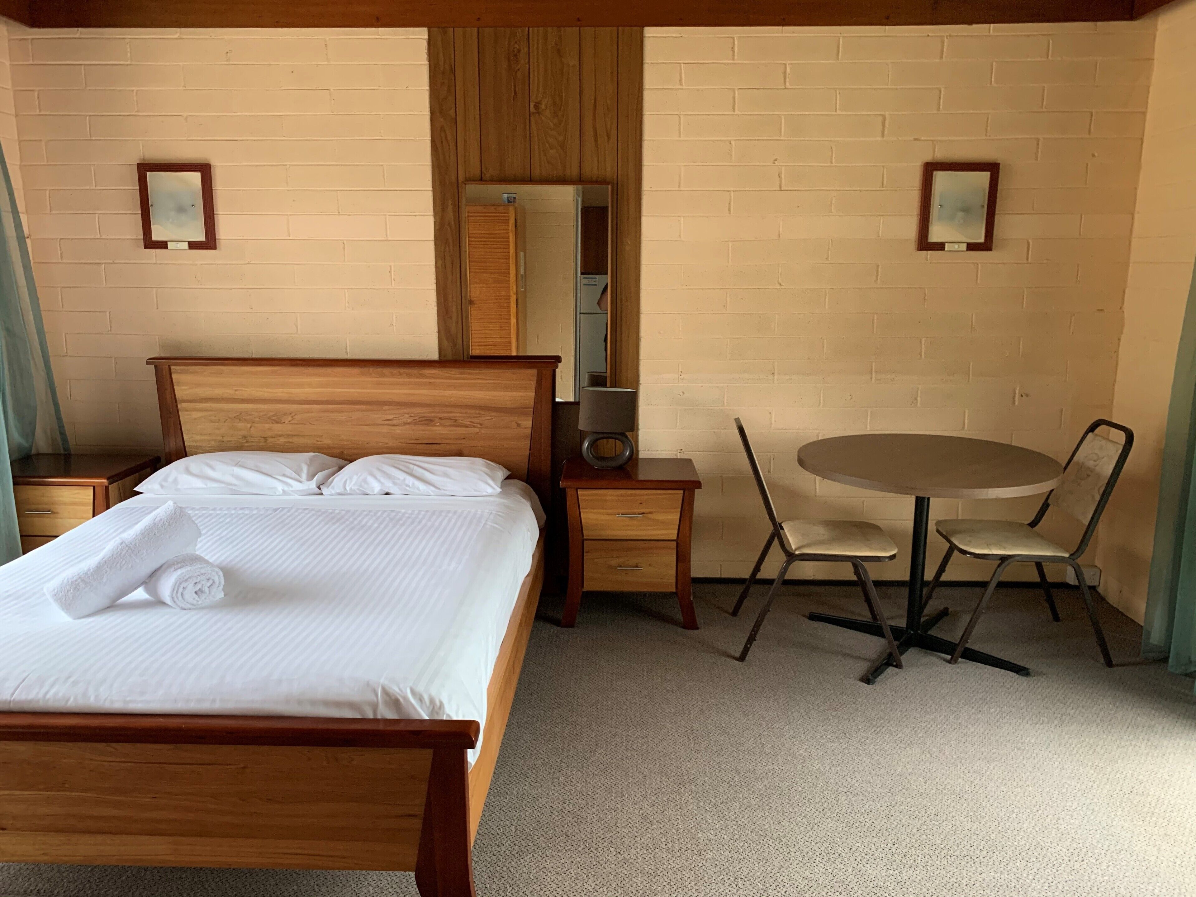 Ein Hotelzimmer mit einem Holzbett, einem runden Tisch mit zwei Stühlen, einem kleinen Nachttisch und einem Spiegel an der Wand.