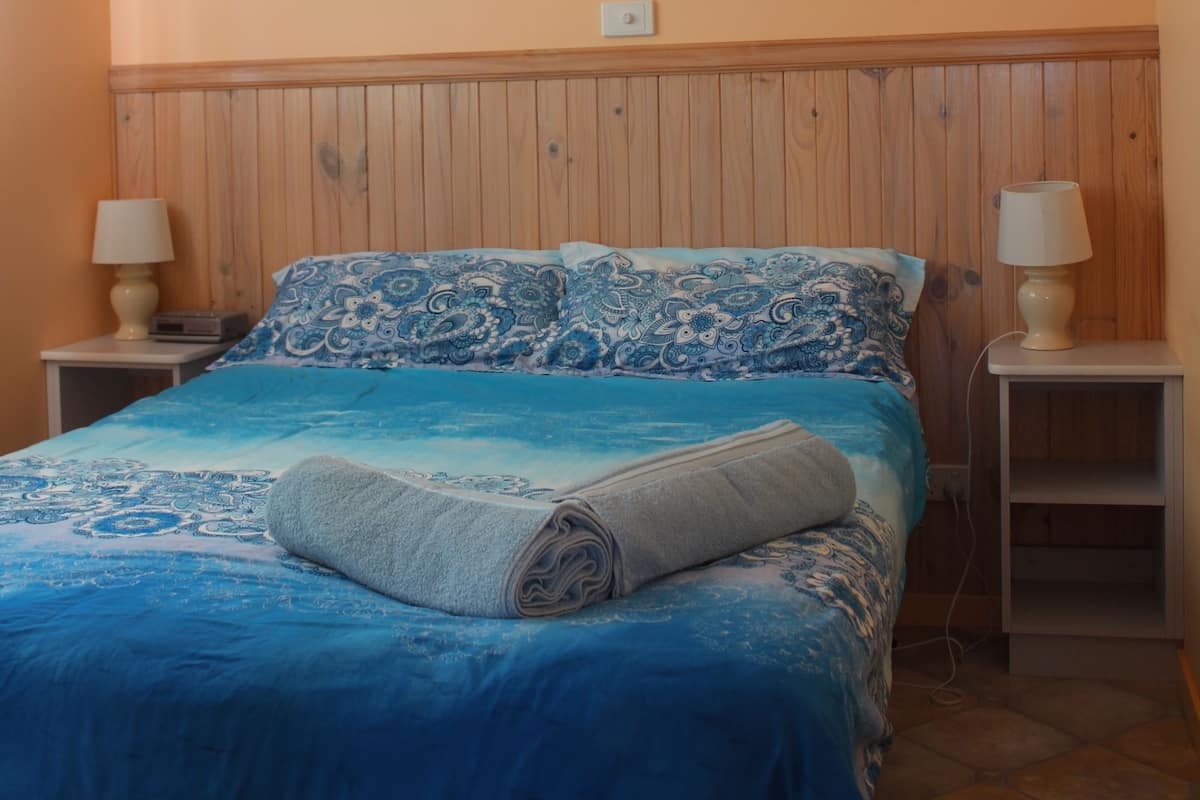 A bedroom with a wooden headboard, a blue bedspread, two bedside tables with lamps, and a framed picture on the wall.