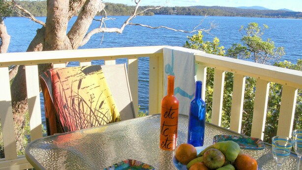 Una mesa de vidrio con un frutero, dos botellas y un vaso en una terraza con vista a un lago.
