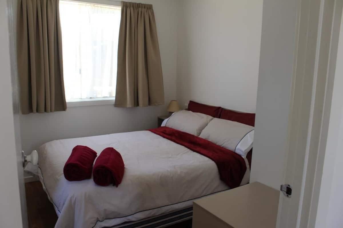A neatly made bed with white and red linens, a bedside lamp, and a window with curtains.