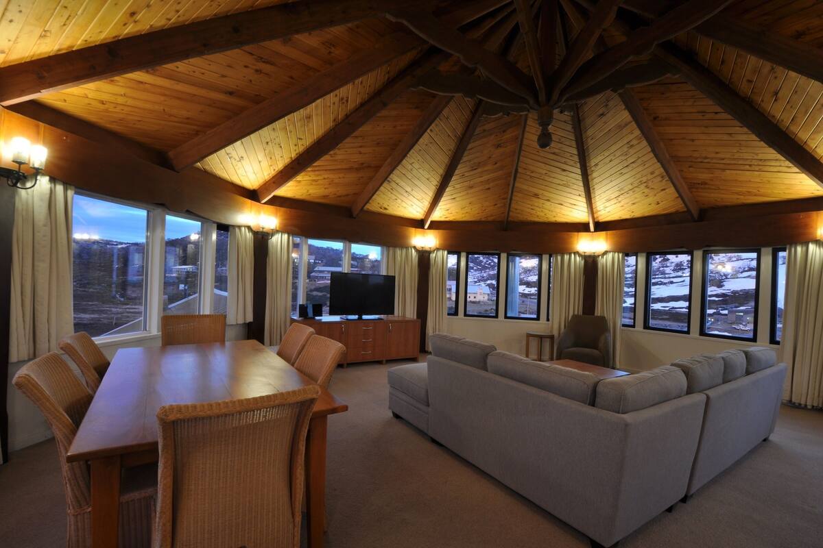 A spacious living room with a high wooden ceiling, a large sofa, a dining table with chairs, and a television.
