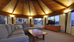 A spacious living room with a large sectional sofa, a wooden coffee table, and a flat-screen TV. The room features a high wooden ceiling and large windows with curtains. - Marritz Hotel (Perisher Valley)