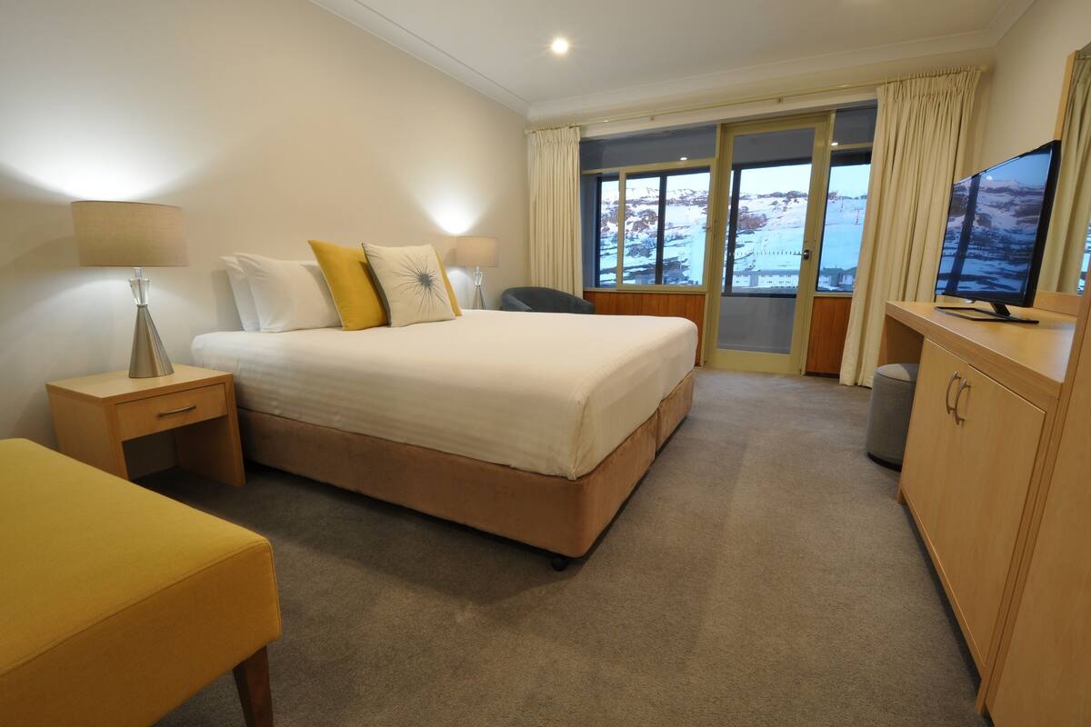A hotel room with a large bed, a desk, a chair, a television, and a view of a snowy landscape through the window.
