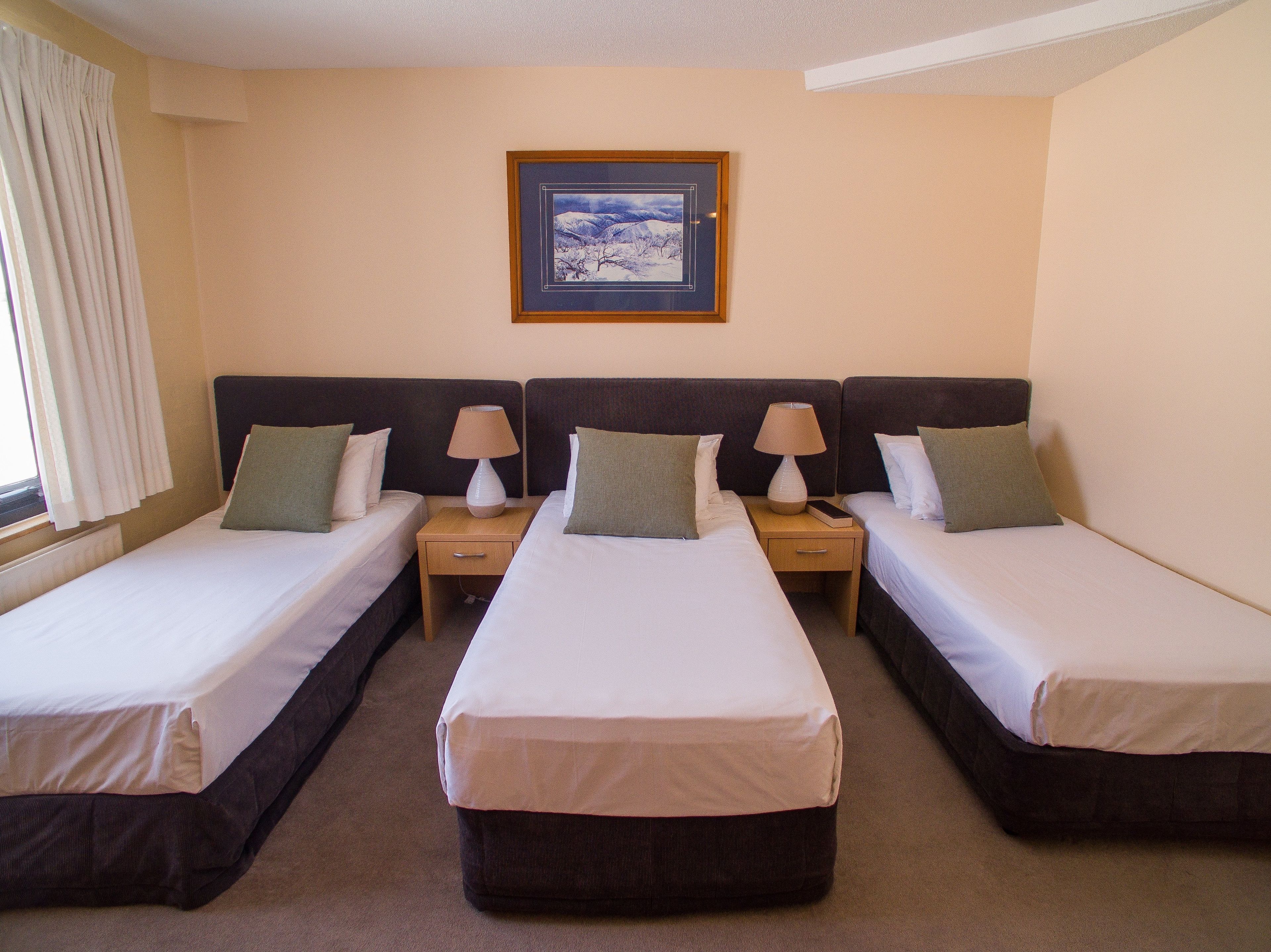 Habitación de hotel con tres camas individuales, cada una con una almohada y una mesita de noche con lámpara y un libro.