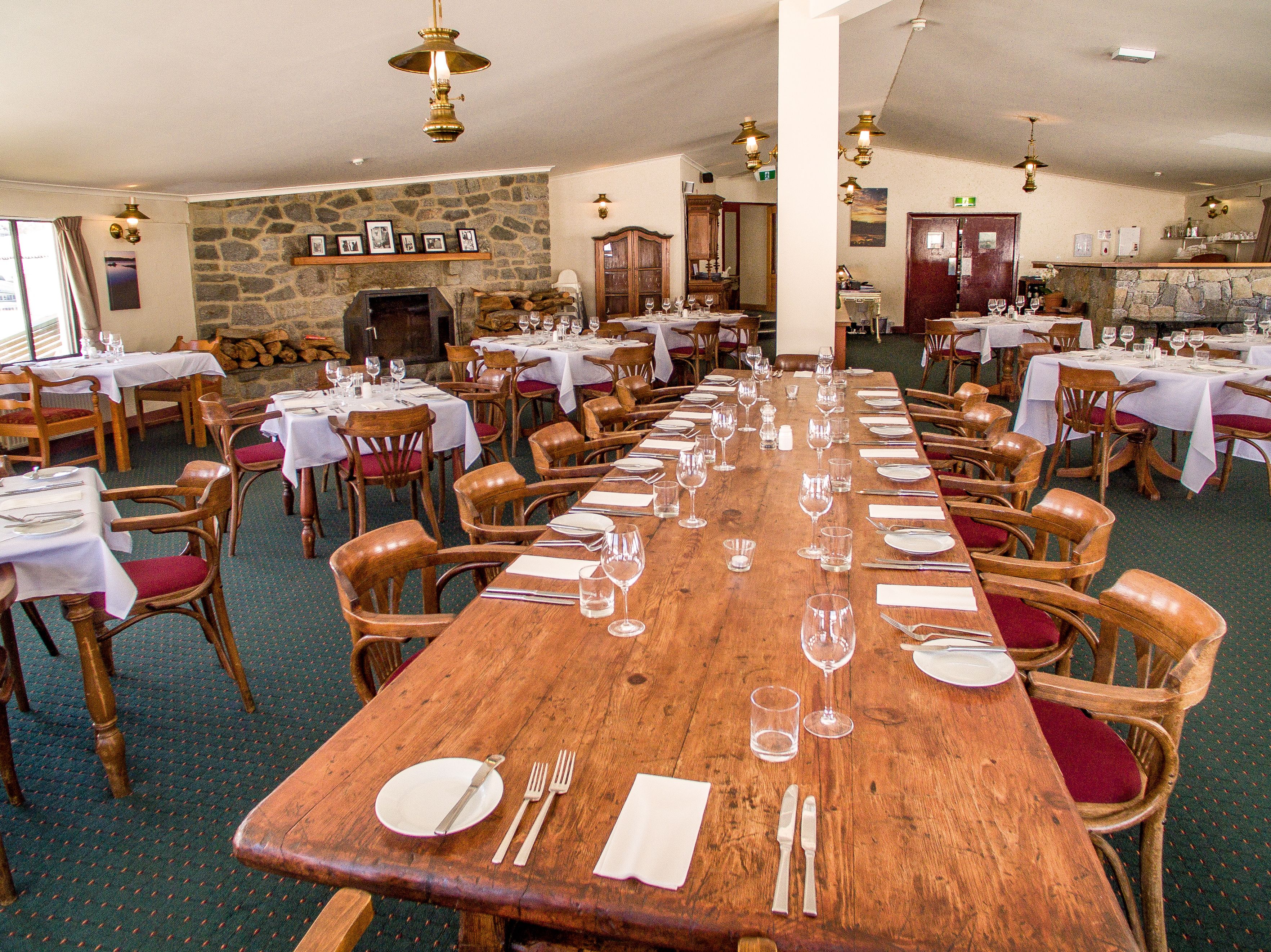 A large dining hall with multiple tables set for a meal, wooden chairs, and a stone wall with framed pictures.