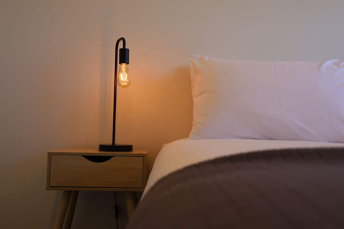 Una mesita de noche con lámpara, un cajón y una cama con ropa de cama blanca.