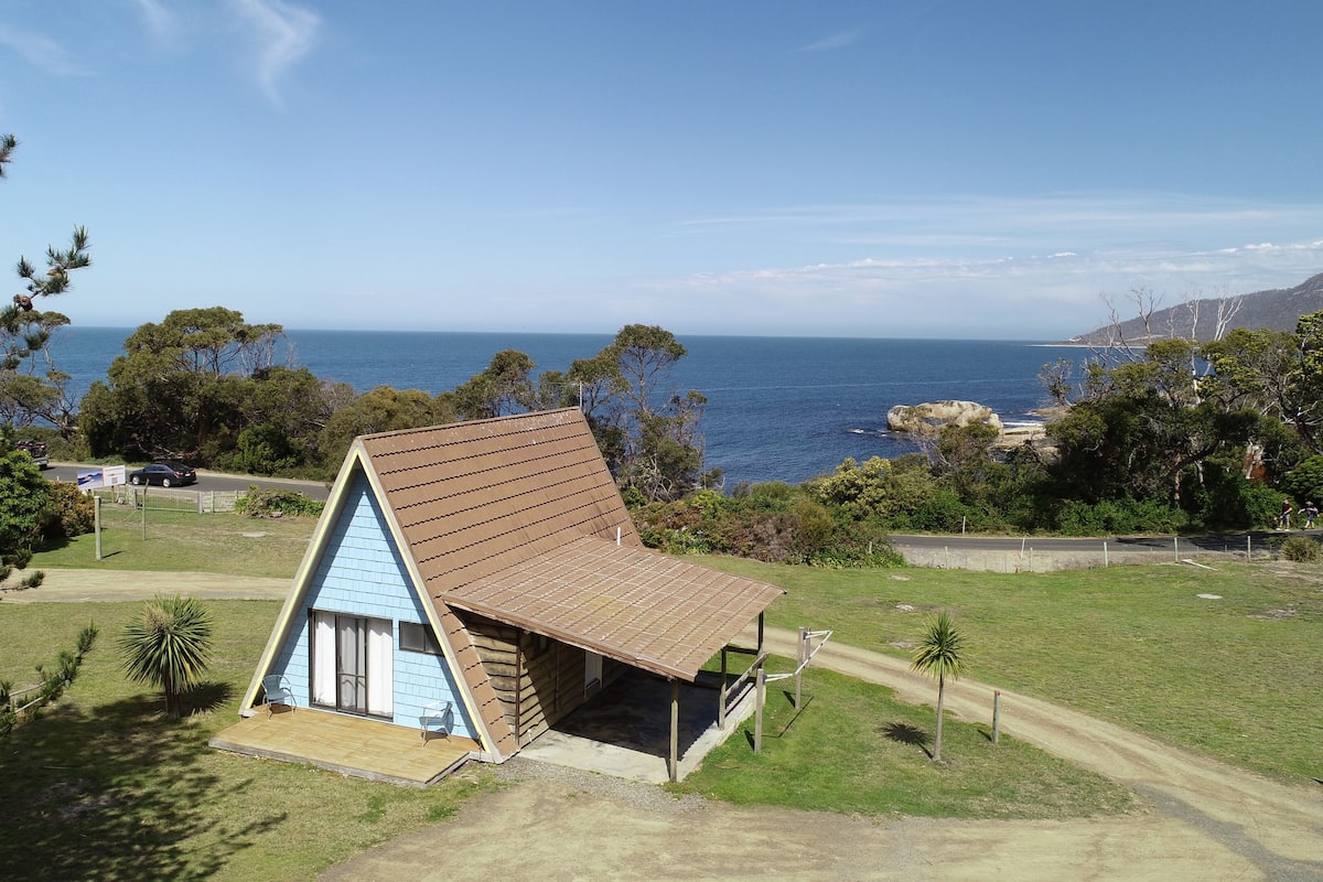 Una pequeña casa de madera con forma triangular y techo inclinado, ubicada en un área cubierta de pasto con vista al océano.