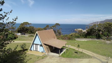 A small, triangular wooden house with a sloped roof, situated on a grassy area with a view of the ocean.
