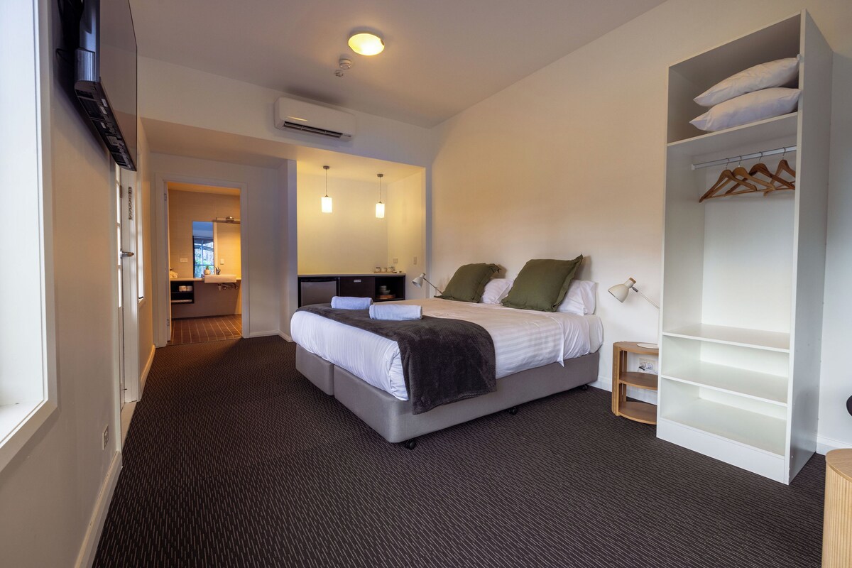 Habitación de hotel con cama, armario, escritorio con silla y baño visible a través de una puerta abierta.