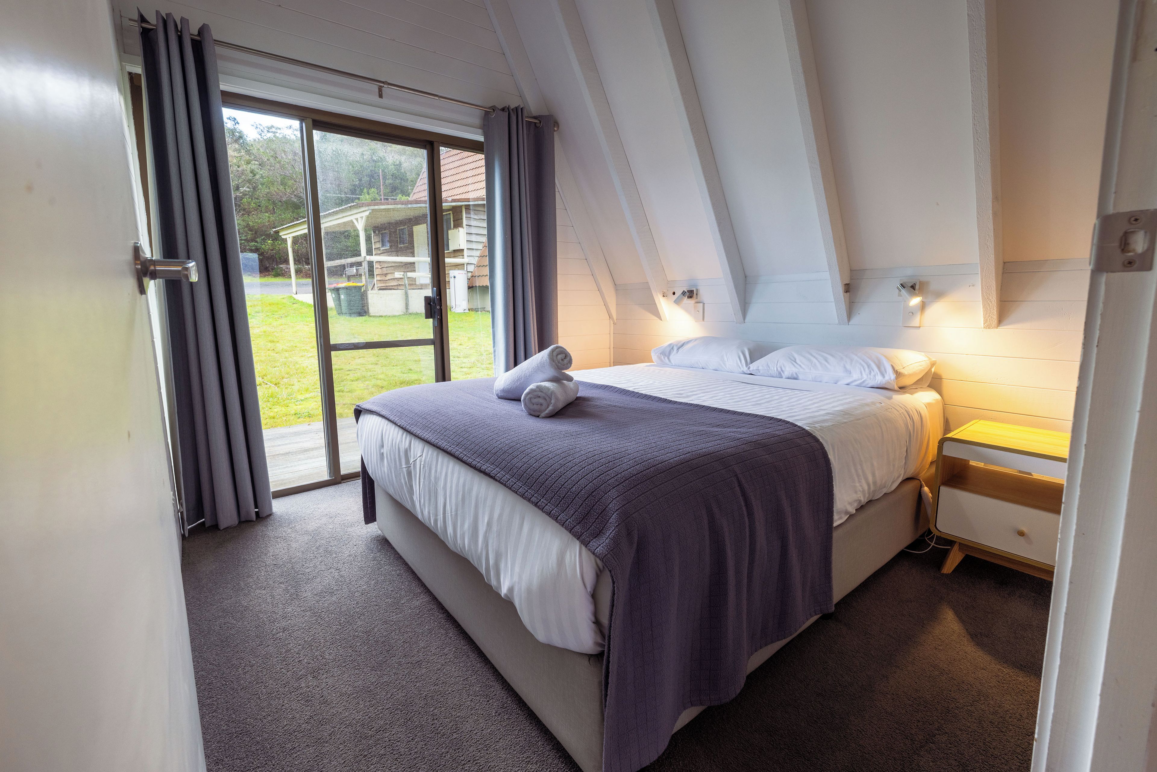 A bedroom with a bed, a nightstand, a lamp, and a view of the outdoors through a sliding door.