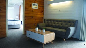 Una habitación con pared de madera, un sofá verde, una mesa de centro de madera con dos plantas pequeñas y una cama al fondo.