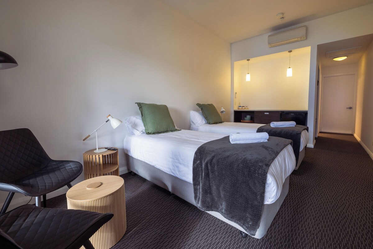 Habitación de hotel con dos camas, una zona de estar con una silla y una mesita, y un baño visible a través de una puerta abierta.