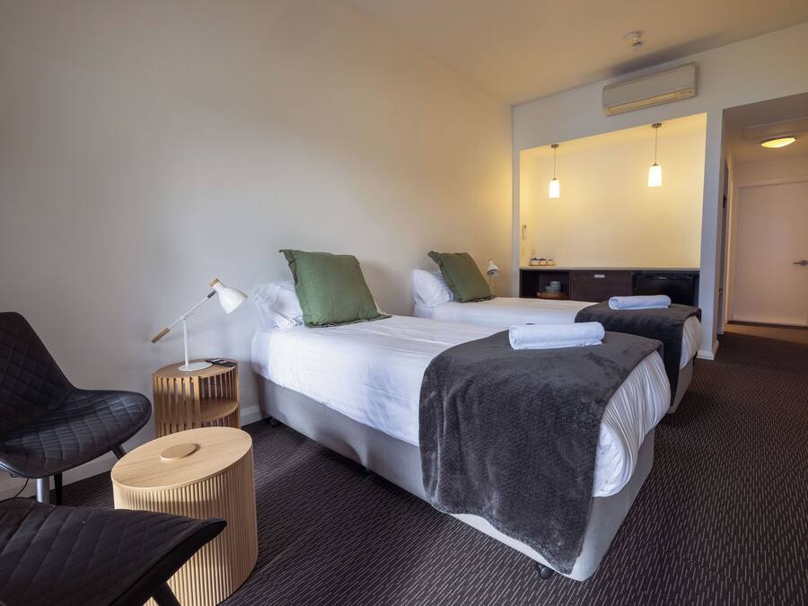 Una camera d'albergo con due letti, un'area salotto con una poltrona e un tavolino, e un bagno parzialmente visibile attraverso una porta aperta.