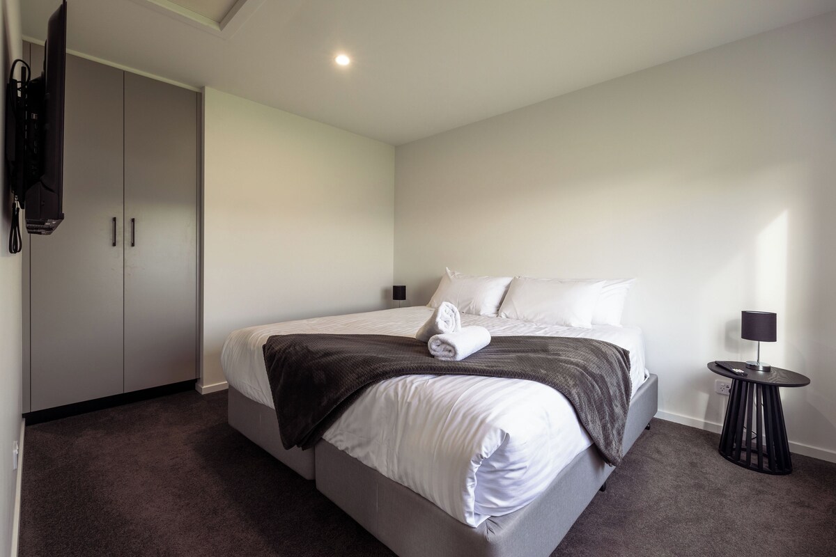 Un dormitorio moderno con cama, televisor montado en la pared y una mesita de noche con lámpara.
