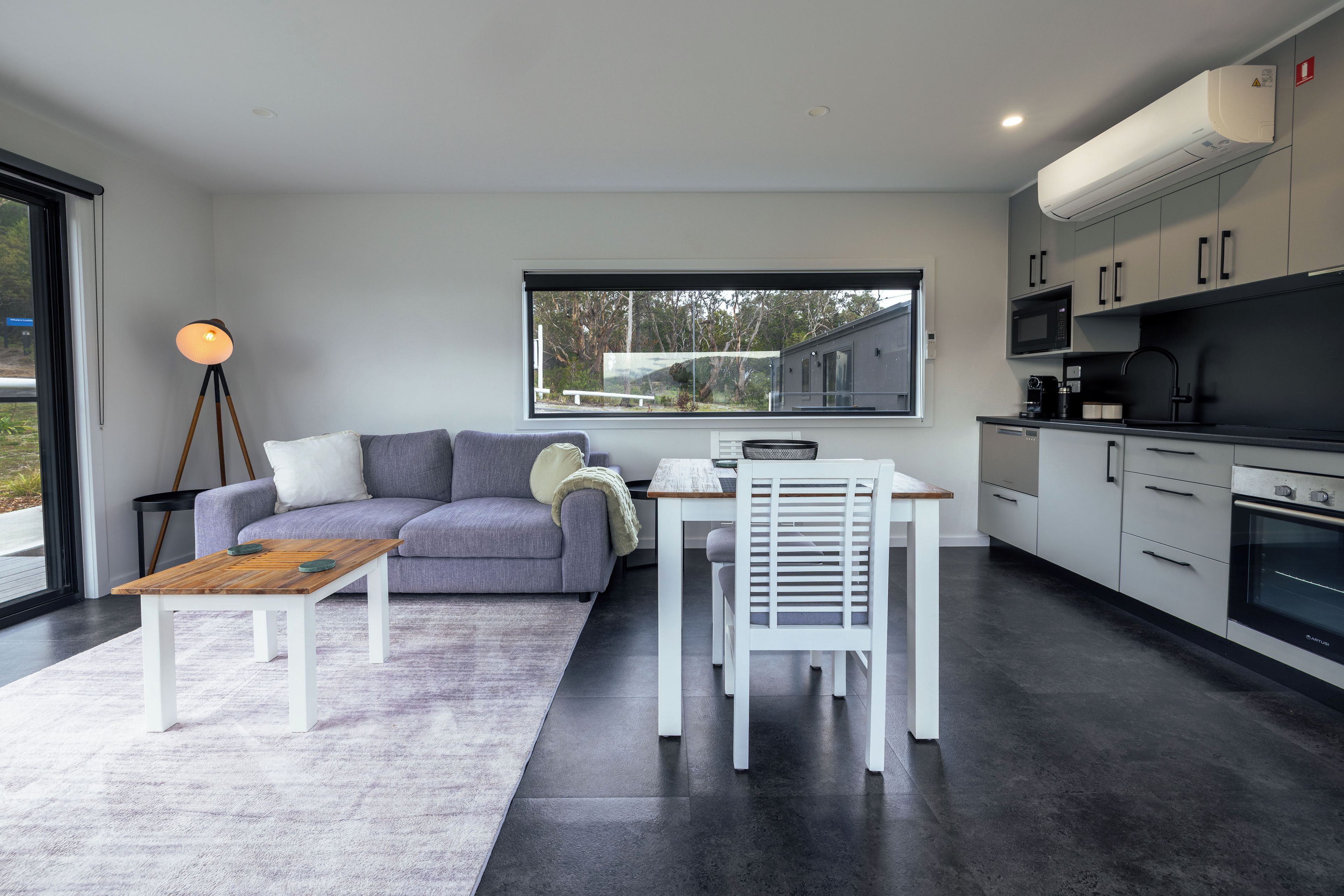 A modern kitchen with white cabinetry, a dining area with a white table and chair, and a grey sofa with cushions.