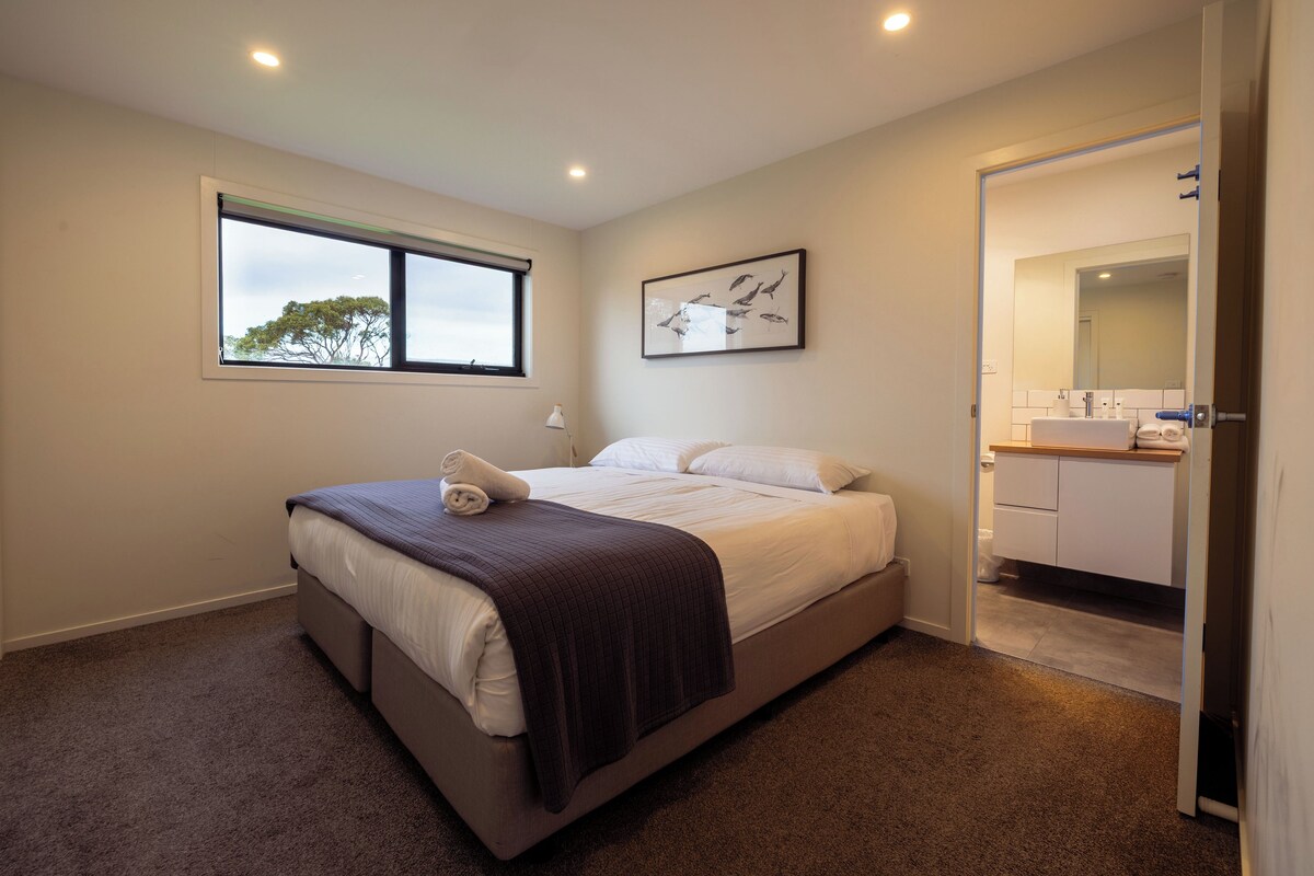 Un dormitorio con cama, ventana, un cuadro enmarcado y un baño visible a través de una puerta abierta.