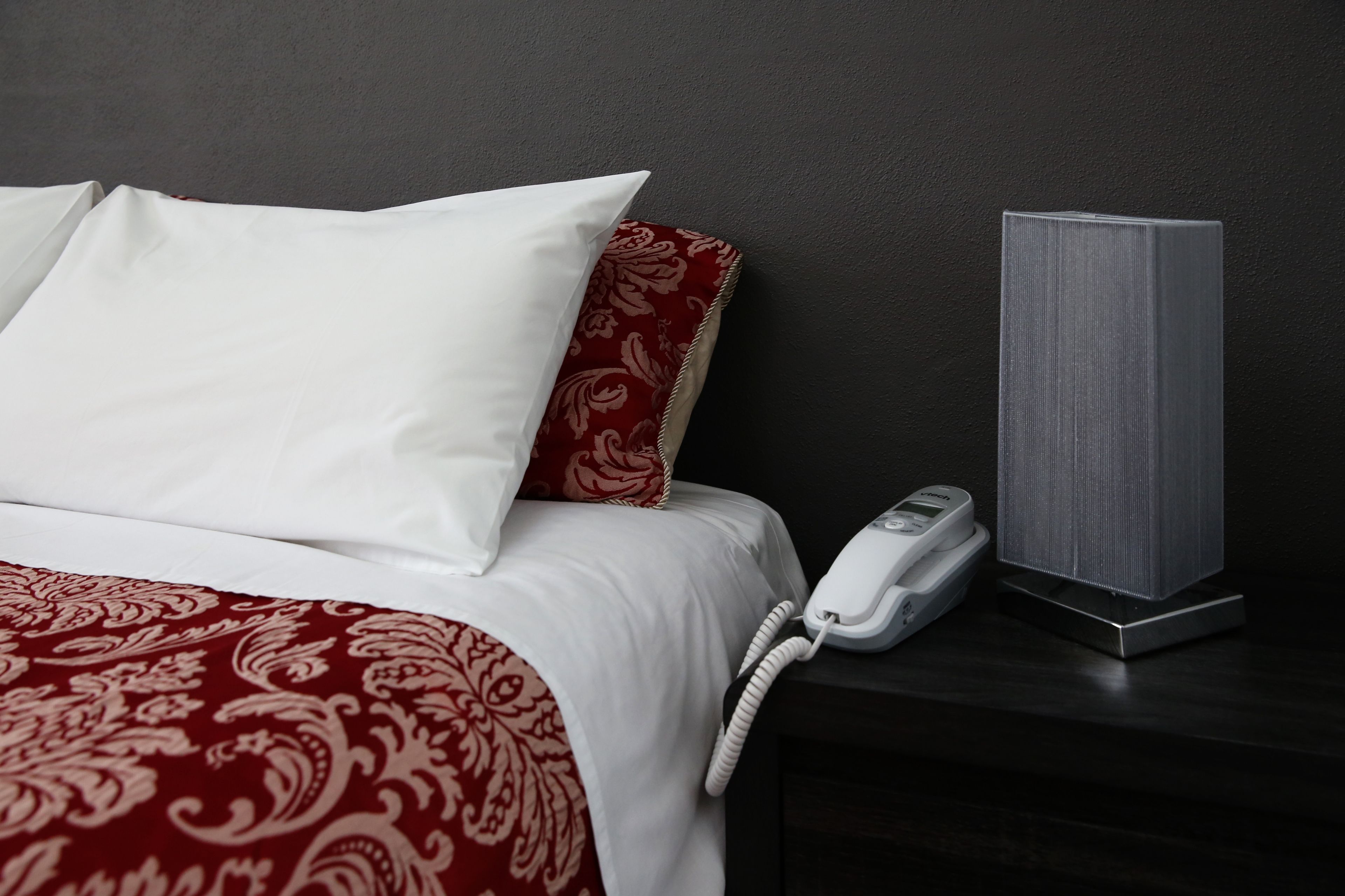 Una cama con almohadas blancas y rojas, un teléfono blanco en la mesita de noche y un altavoz gris sobre una superficie oscura.