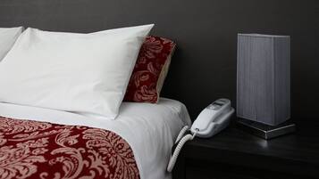 Una cama con almohadas blancas y rojas, un teléfono blanco en la mesita de noche y un altavoz gris sobre una superficie oscura.