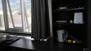 Une pièce avec une fenêtre, un bureau, une machine à café et une étagère contenant de la vaisselle et une horloge.