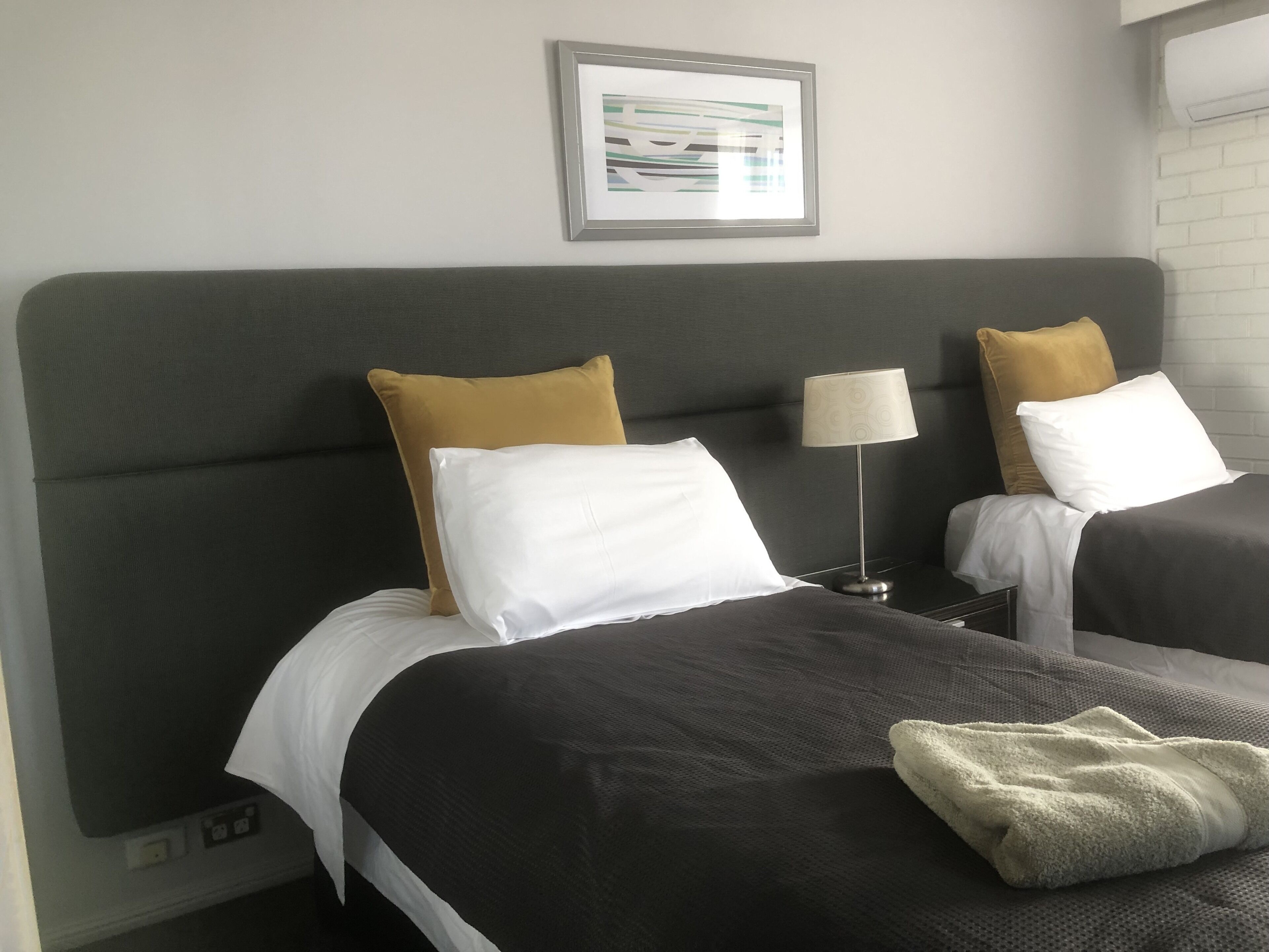 Una habitación de hotel con dos camas, cada una con cabecero gris, almohadas blancas y mostaza, una mesita de noche con lámpara y un cuadro en la pared.