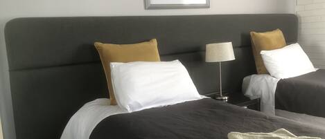 Una habitación de hotel con dos camas, cada una con cabecero gris, almohadas blancas y mostaza, una mesita de noche con lámpara y un cuadro en la pared.