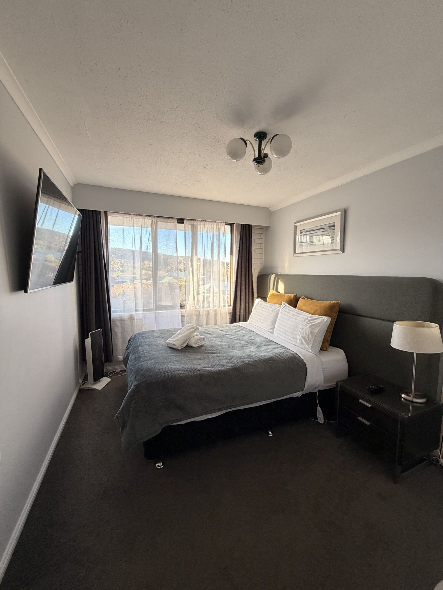 A hotel room with a bed, a bedside table, a lamp, a window with curtains, and a ceiling fan.