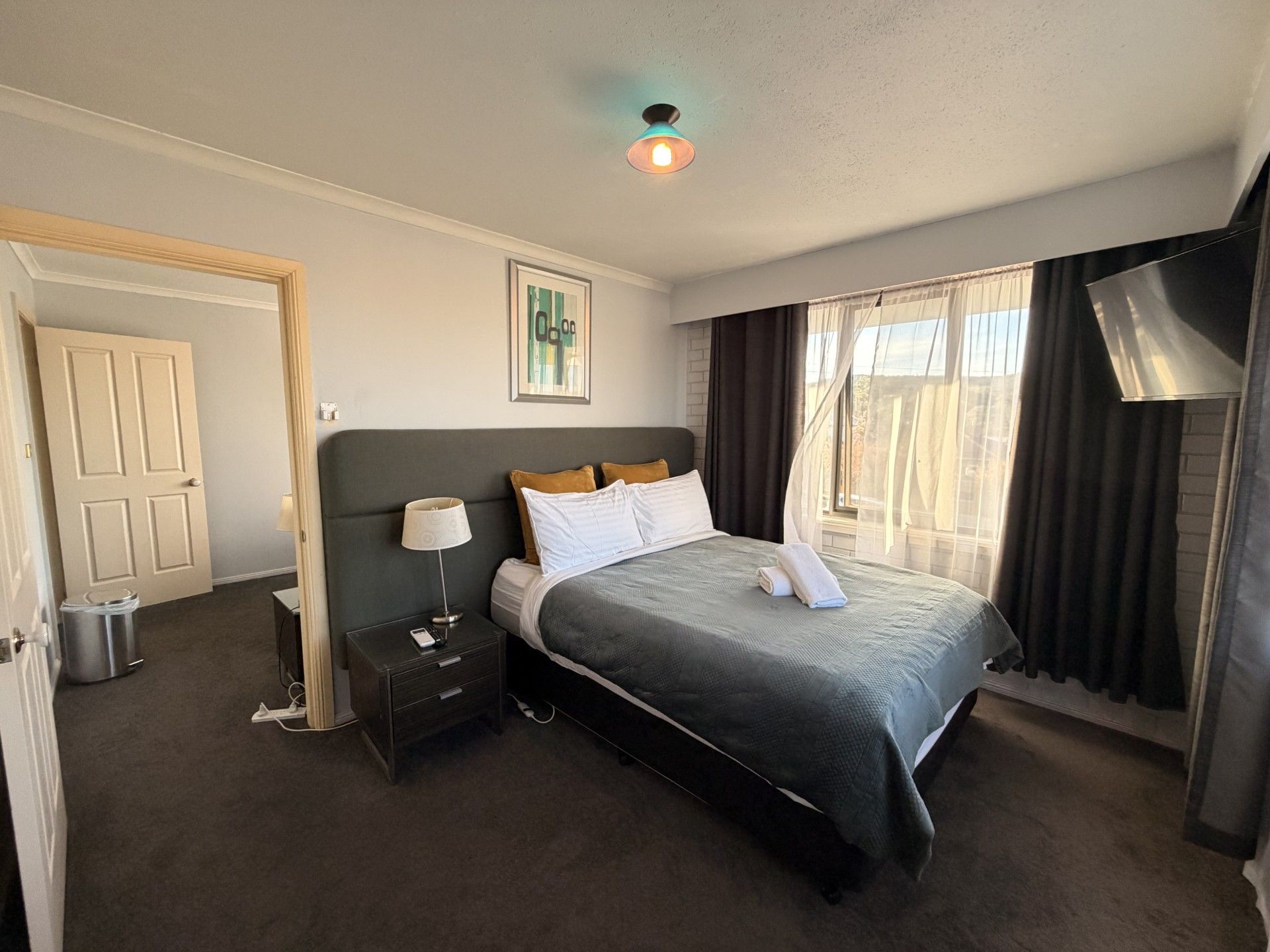 A hotel room with a bed, bedside table, lamp, and a window with curtains.