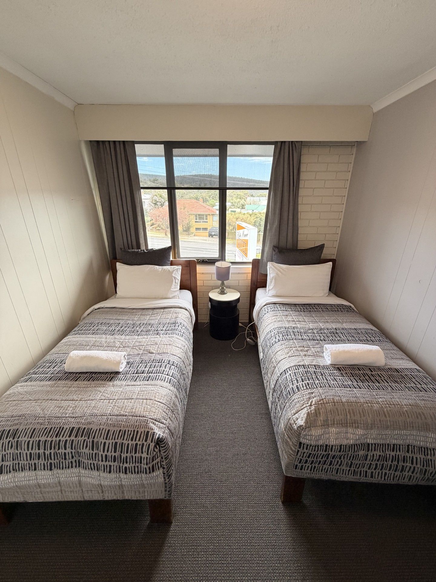 A twin bedroom with two beds, a nightstand, and a window with a view of buildings.