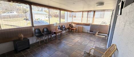 A covered patio area with stone flooring, a seating arrangement including chairs and a table, and large windows providing views of the outside.