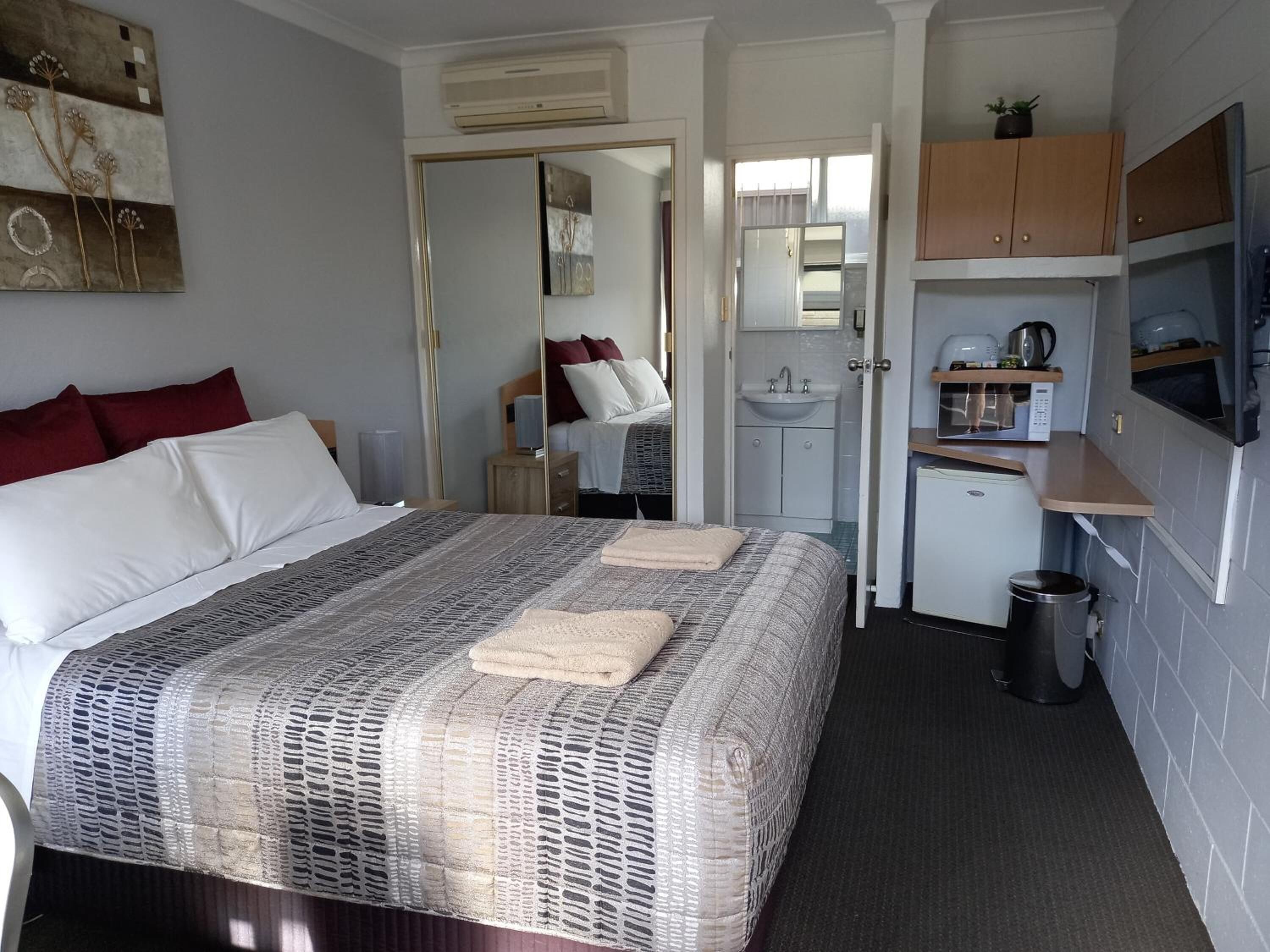 A compact hotel room with a bed, a small kitchenette, a bathroom, and a mirror.
