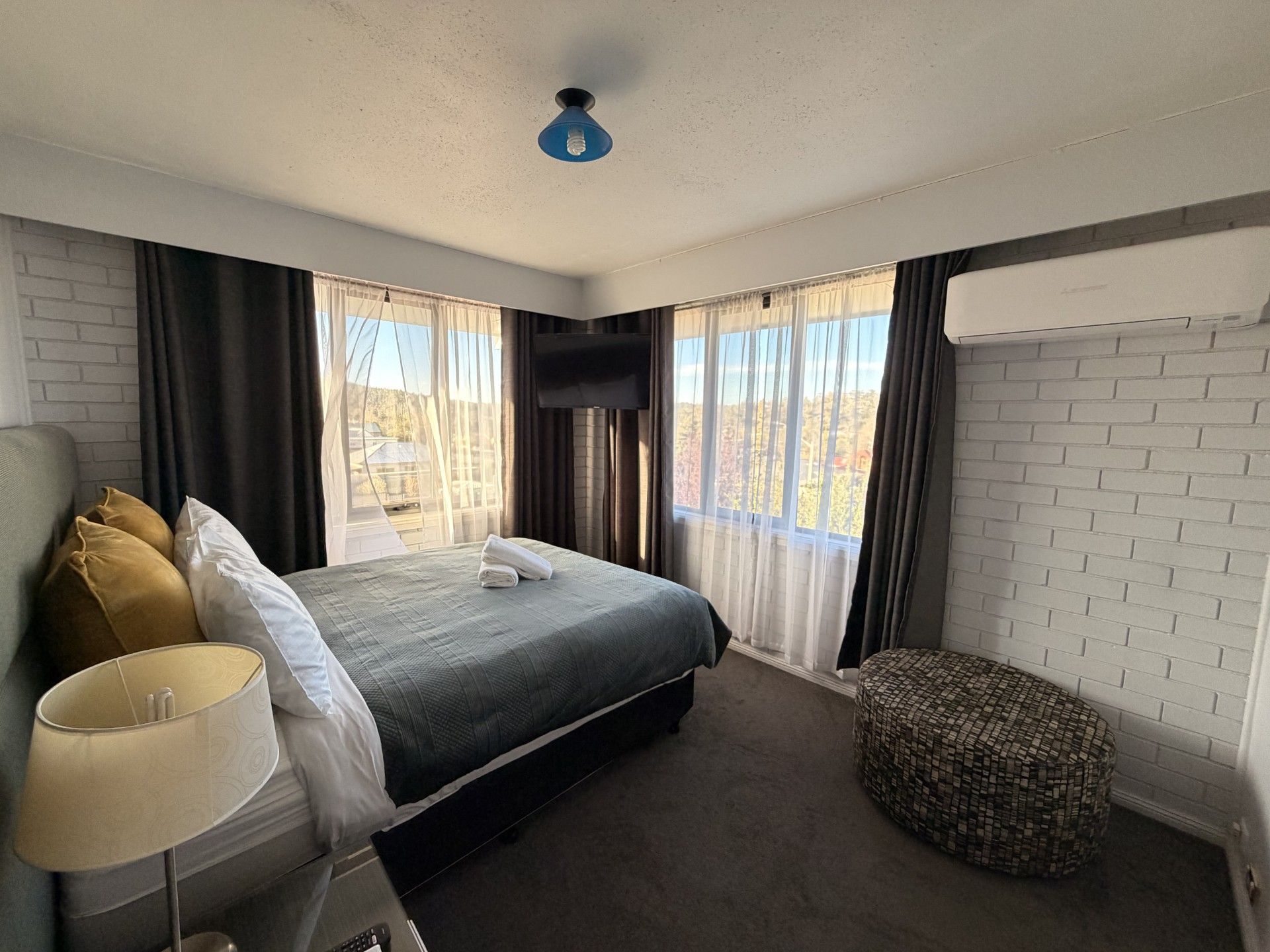 A hotel room with a bed, a bedside table with a lamp, a TV mounted on the wall, and a view of the outdoors.