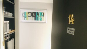 Un couloir avec une porte noire portant le numéro « 14 » et un panneau indiquant « INTERDICTION DE FUMER ». On aperçoit un four à micro-ondes et des étagères remplies d’objets au fond.