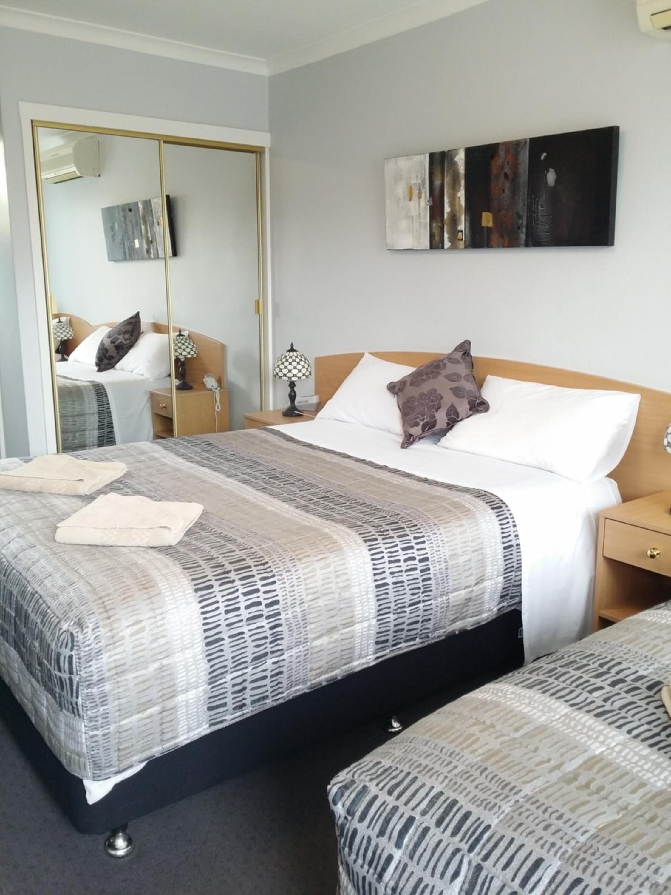 A hotel room with a large bed, a smaller bed, a wardrobe, a bedside table with a lamp, and a painting on the wall.
