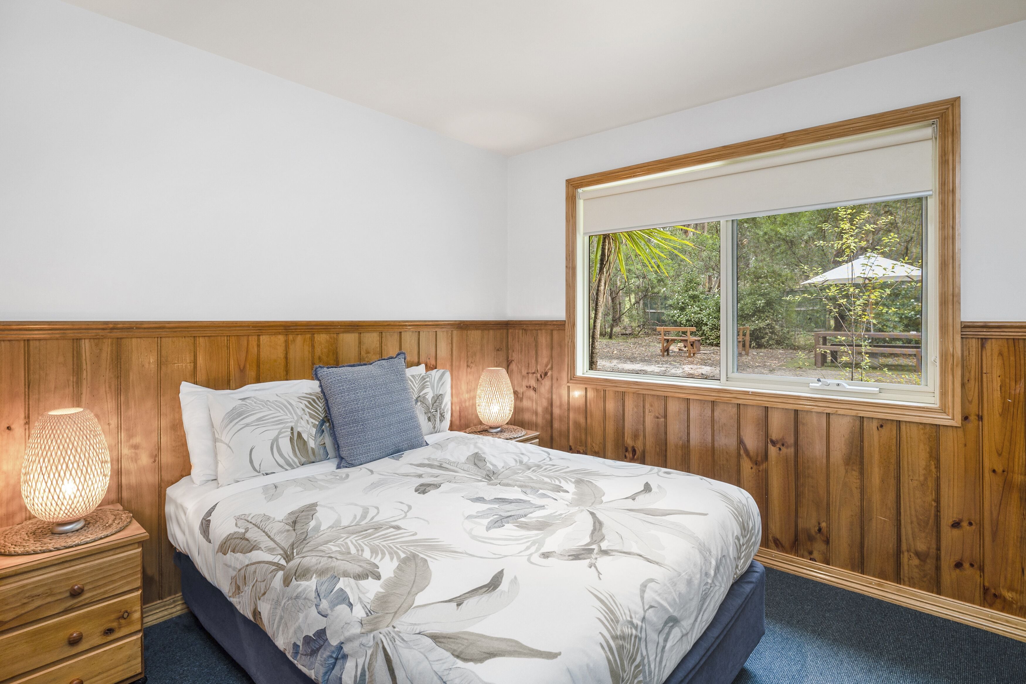 A bedroom with a large bed, wooden headboard, a window with a view of the outdoors, and a nightstand with a lamp.