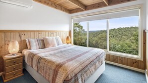 Un dormitorio con un ventanal grande, vigas de madera en el techo, una cama con manta estampada y una lámpara de noche.