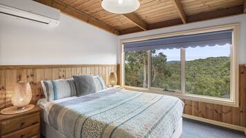 Ein Schlafzimmer mit einem großen Bett, einem Holz-Kopfbrett, einer Nachttischlampe, einem Fenster mit Blick auf Bäume und einer Klimaanlage.