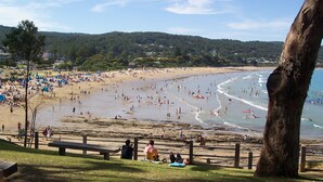 Una playa con gente nadando y tomando el sol, una orilla arenosa y una colina boscosa al fondo.