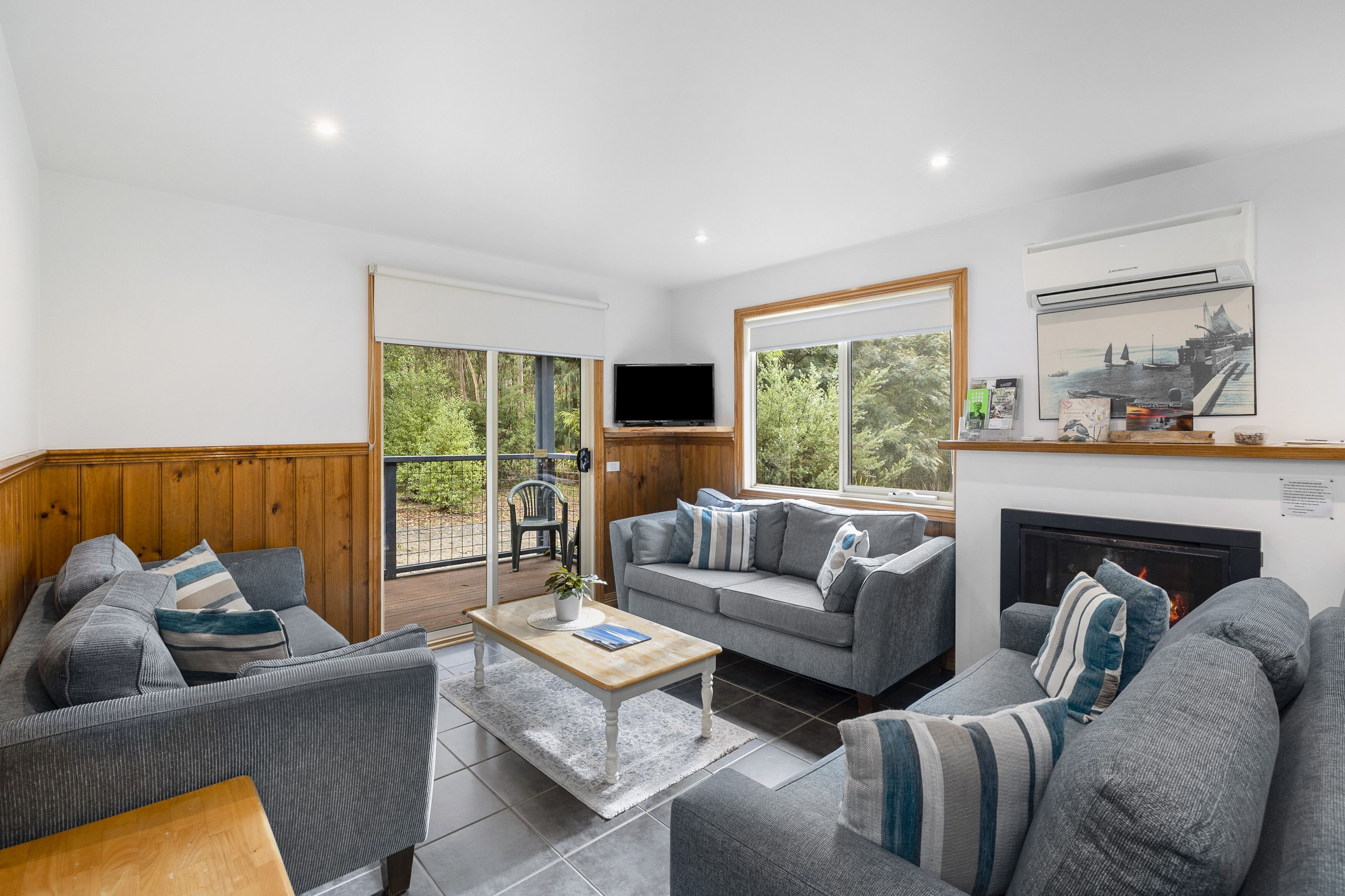 A modern living room with a fireplace, grey sofas, a wooden coffee table, and large windows offering a view of greenery.