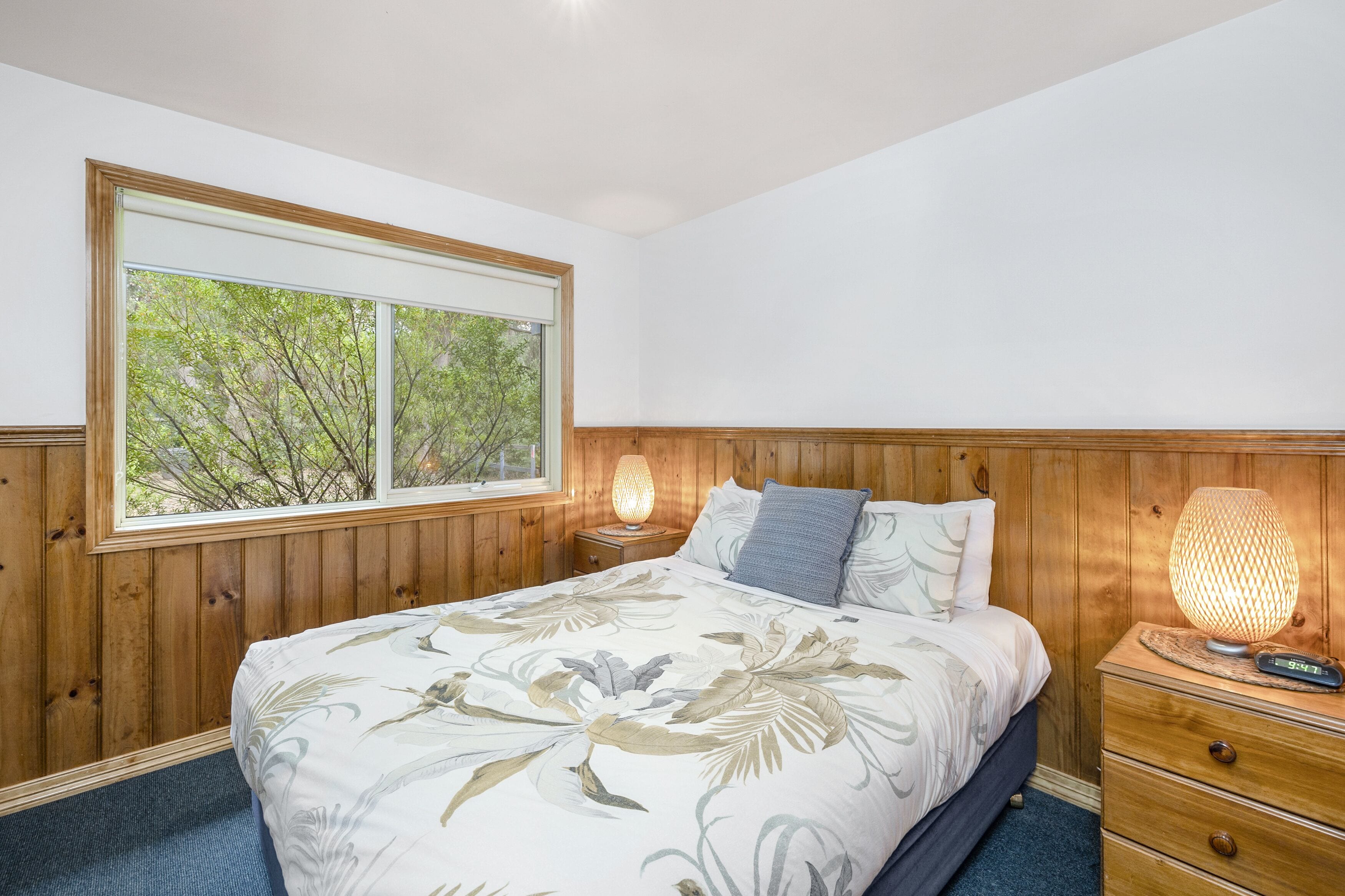 A bedroom with a large window, wooden paneling, a bed with floral bedding, two bedside lamps, and a nightstand with a digital clock.