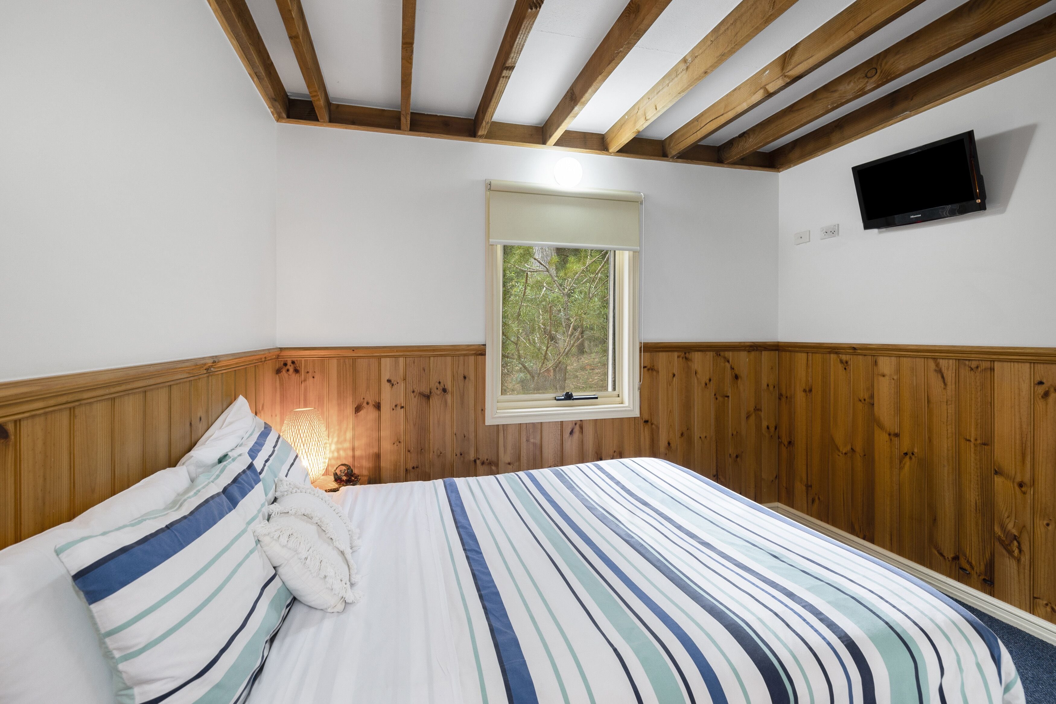 A bedroom with a wooden headboard, a bed with striped bedding, a window with a view of trees, and a mounted TV on the wall.