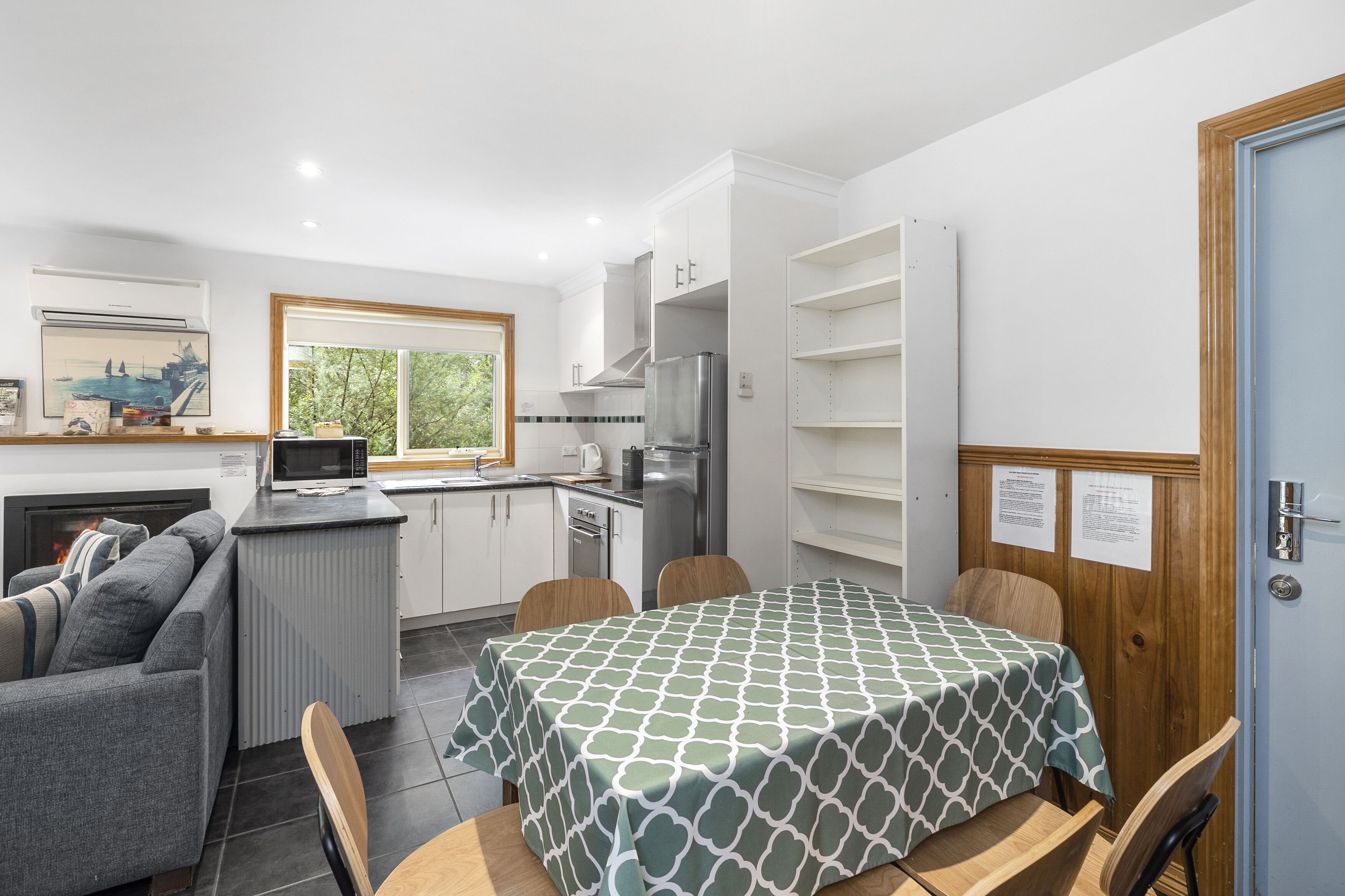 A modern kitchen with white cabinets, a stainless steel refrigerator, and a microwave. There is a dining table with a green patterned tablecloth.